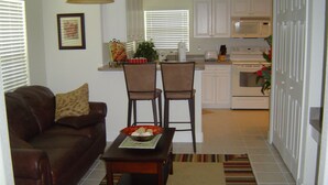 A living room with a brown leather sofa, two chairs, a dining table with a bowl of snacks, and a kitchen in the background with white cabinets and appliances. - Baytree Vacation Villa-- (Rotonda Lakes)