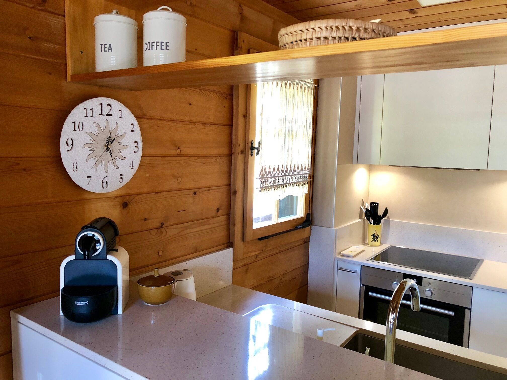 Une cuisine avec des murs en bois, une horloge ronde, une étagère pour les boîtes à café et à thé, une machine à café et un évier doté d'un robinet moderne.