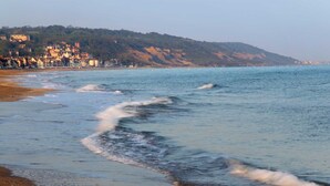 A coastal scene with waves approaching the shore, a sandy beach, and a hillside with buildings in the background.