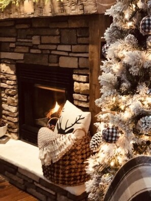 Ein Kamin aus Stein mit brennendem Feuer, ein geschmückter Weihnachtsbaum und ein kariertes Kissen auf dem Sims.