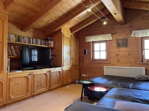 Une chambre confortable, aménagée dans le style d’un chalet, avec des murs et un plafond en bois, un téléviseur posé sur un meuble en bois, une bibliothèque remplie de livres, une fenêtre avec des rideaux et un lit recouvert d’une couverture.