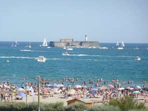 A crowded beach with people sunbathing and swimming, boats on the water, and a lighthouse in the background.