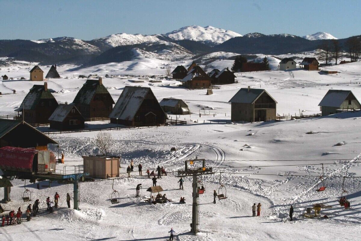 Un village de montagne enneigé, avec ses maisons traditionnelles en bois, un télésiège et une vue dégagée sur les montagnes recouvertes de neige en arrière-plan.