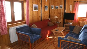 Un accogliente interno di una baita con pareti in legno, un divano rosso, poltrone blu, un tavolino in legno e un televisore.