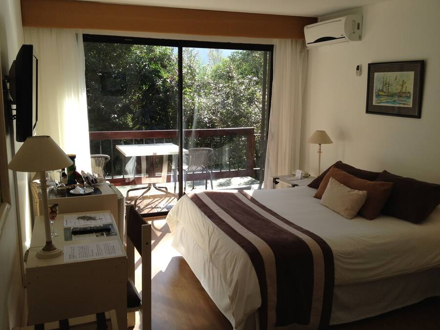 A hotel room with a bed, a desk with a lamp, a chair, and a view of the outdoors through a large window.