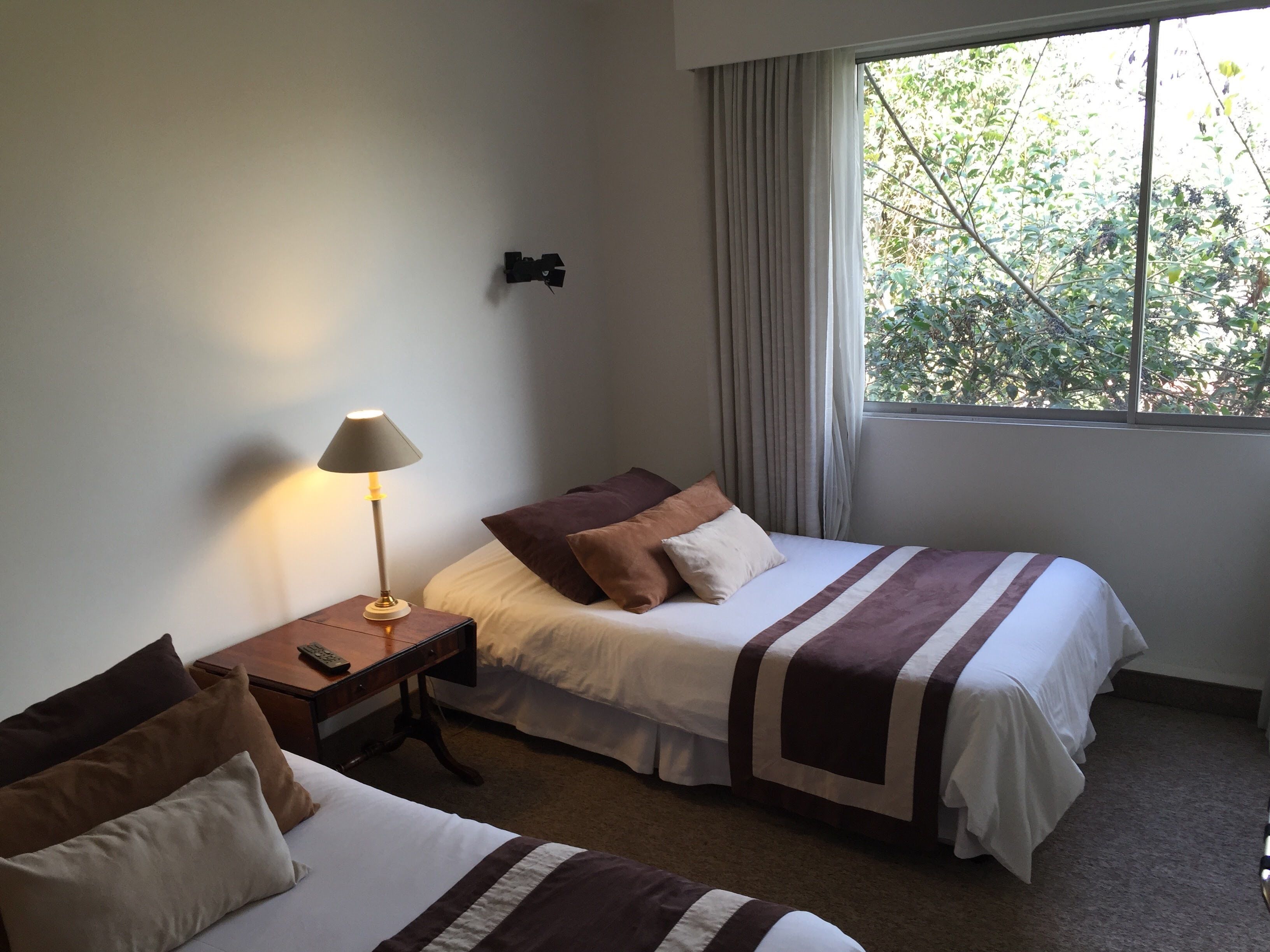 Quarto de hotel com duas camas, uma mesa de cabeceira com abajur, uma janela com cortinas e vista para árvores.