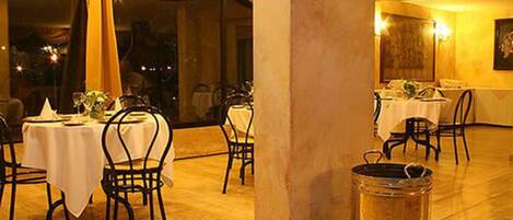 Um restaurante com várias mesas e cadeiras, cortinas penduradas e vista para a noite lá fora.