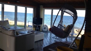 Habitación con sofá blanco, una silla colgante y televisor. Cuenta con ventanas amplias con vista al mar.