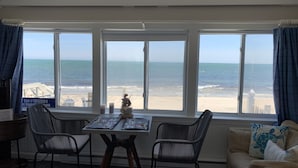 Una habitación con vista a la playa, que incluye una mesa con sillas, un sofá y un cartel con la palabra 'Welcome' sobre las ventanas.