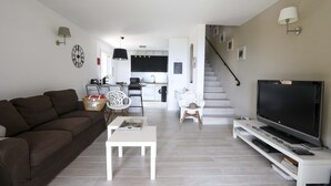 Una sala de estar moderna con un sofá marrón, una mesa de centro blanca y un televisor sobre un mueble blanco. Se ve una escalera que lleva a una cocina al fondo.