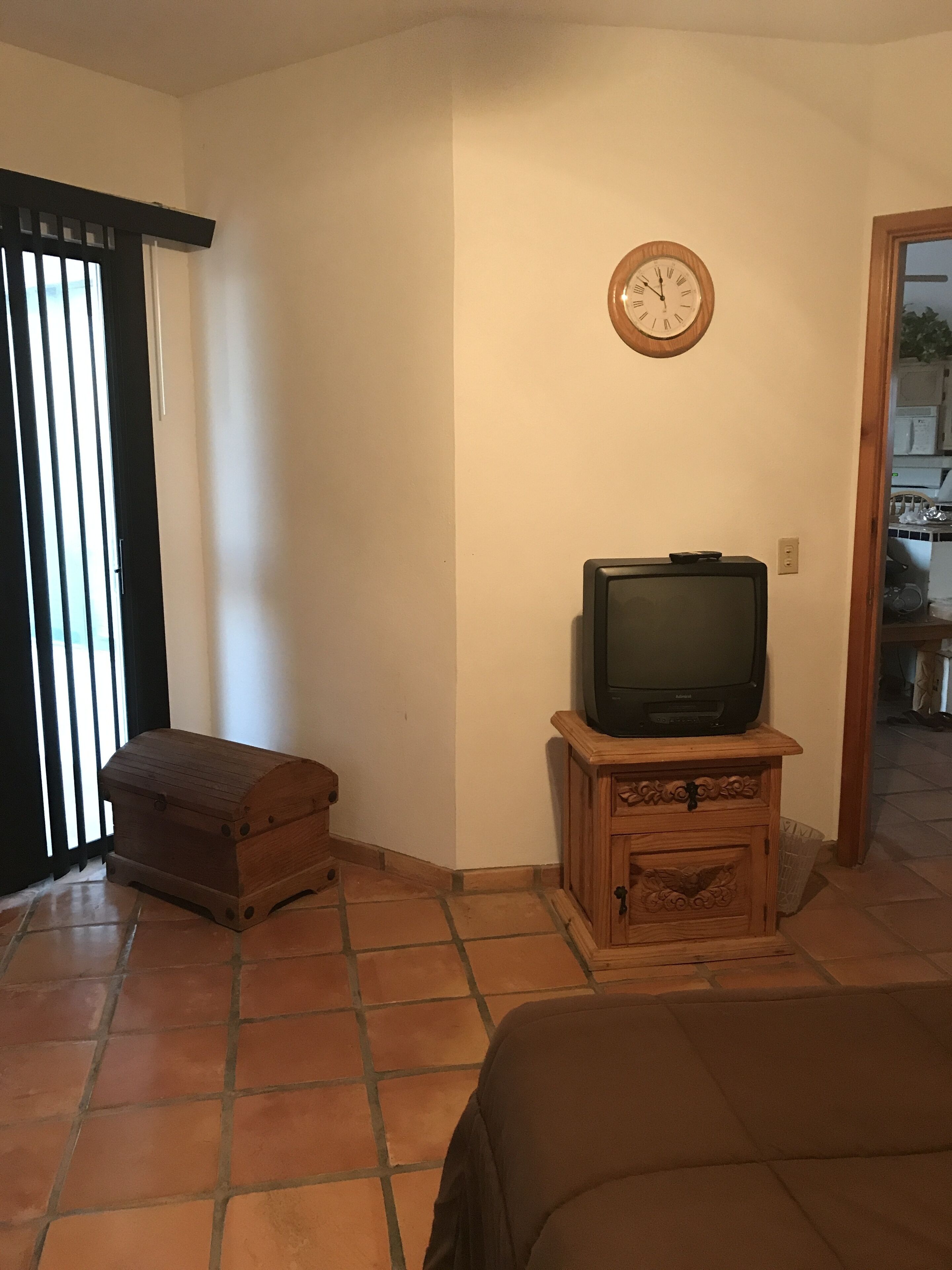 A living room with a television on a wooden stand, a wooden chest, and a clock on the wall.