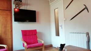 Ett rum med ett träbord, en rosa stol, en tv som är monterad på väggen, ett fönster med persienner och ett element.
