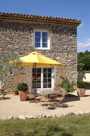 Un bâtiment en pierre avec un toit en tuiles, une fenêtre et un patio agrémenté d’un parasol jaune, de chaises et de plantes en pot.