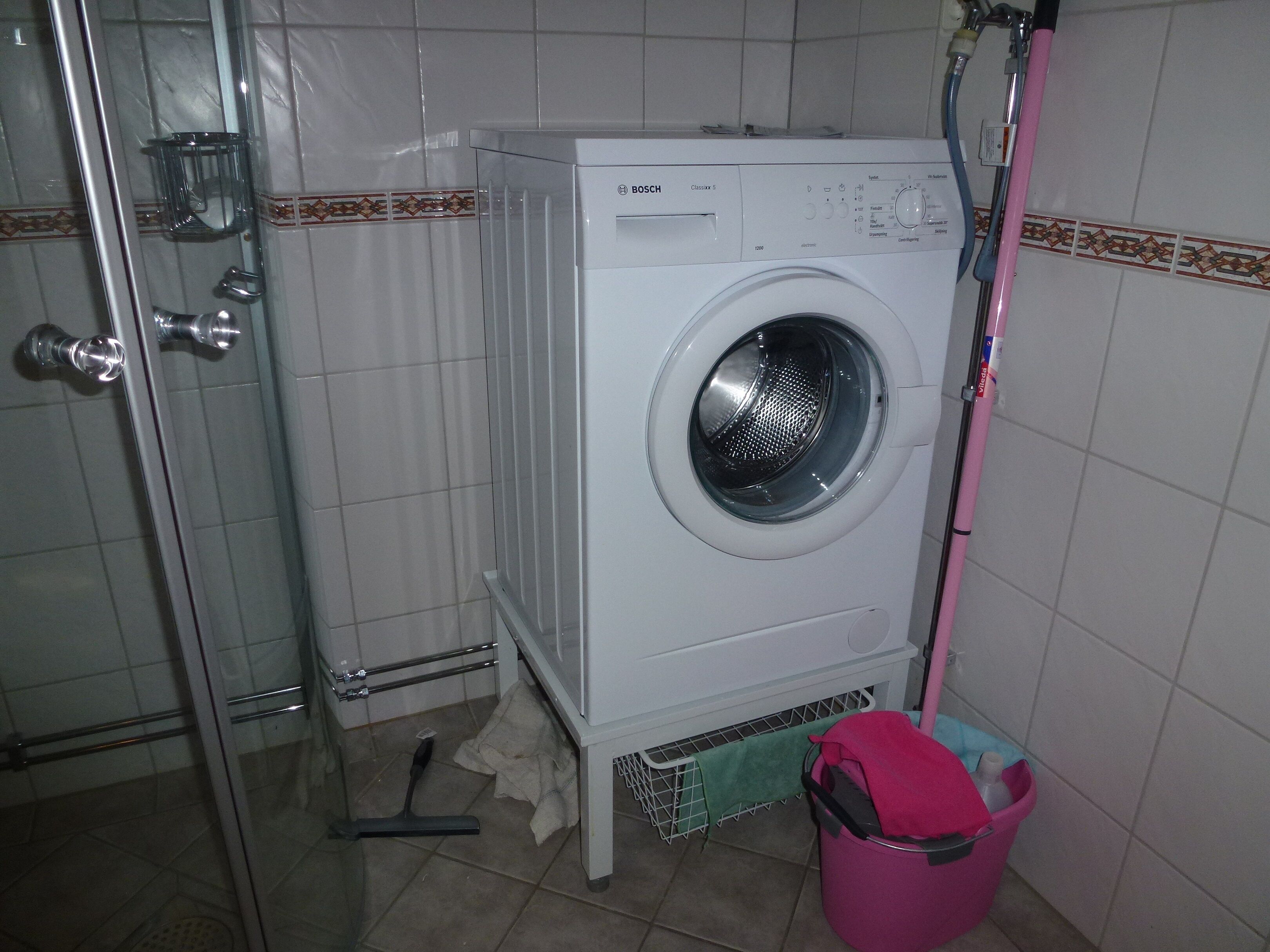 A compact, white Bosch washing machine in a small laundry room with tiled walls, a metal rack, and a pink bucket.