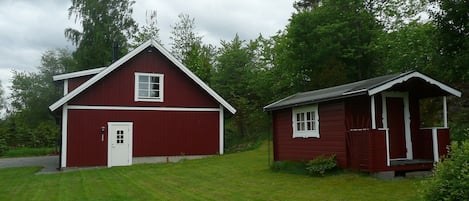 A red barn with a white door and window, a smaller shed with a porch, and a grassy yard with trees in the background.