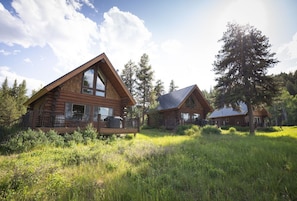 A log cabin with a large deck, surrounded by tall trees and a grassy field.