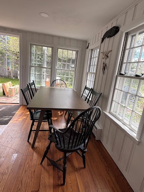 A dining area with a wooden table and chairs, large windows, and a view of the outdoors.