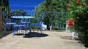 Eine Terrasse mit blauen Metallstühlen und einem Tisch, mit Blick auf ein Gewässer und Berge.