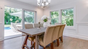 A dining room with a wooden table and beige upholstered chairs, large windows, and a chandelier. - Brand New in the Heart of East Hampton Village with Pool (East Hampton)