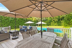 An outdoor deck with a pool, featuring a wooden table, chairs, and umbrellas. - Brand New in the Heart of East Hampton Village with Pool (East Hampton)