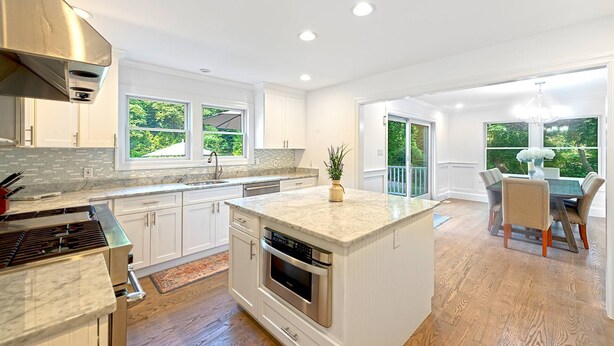 A modern kitchen with white cabinets, a large island, and a dining area with a view of the outdoors. - Brand New in the Heart of East Hampton Village with Pool (East Hampton)