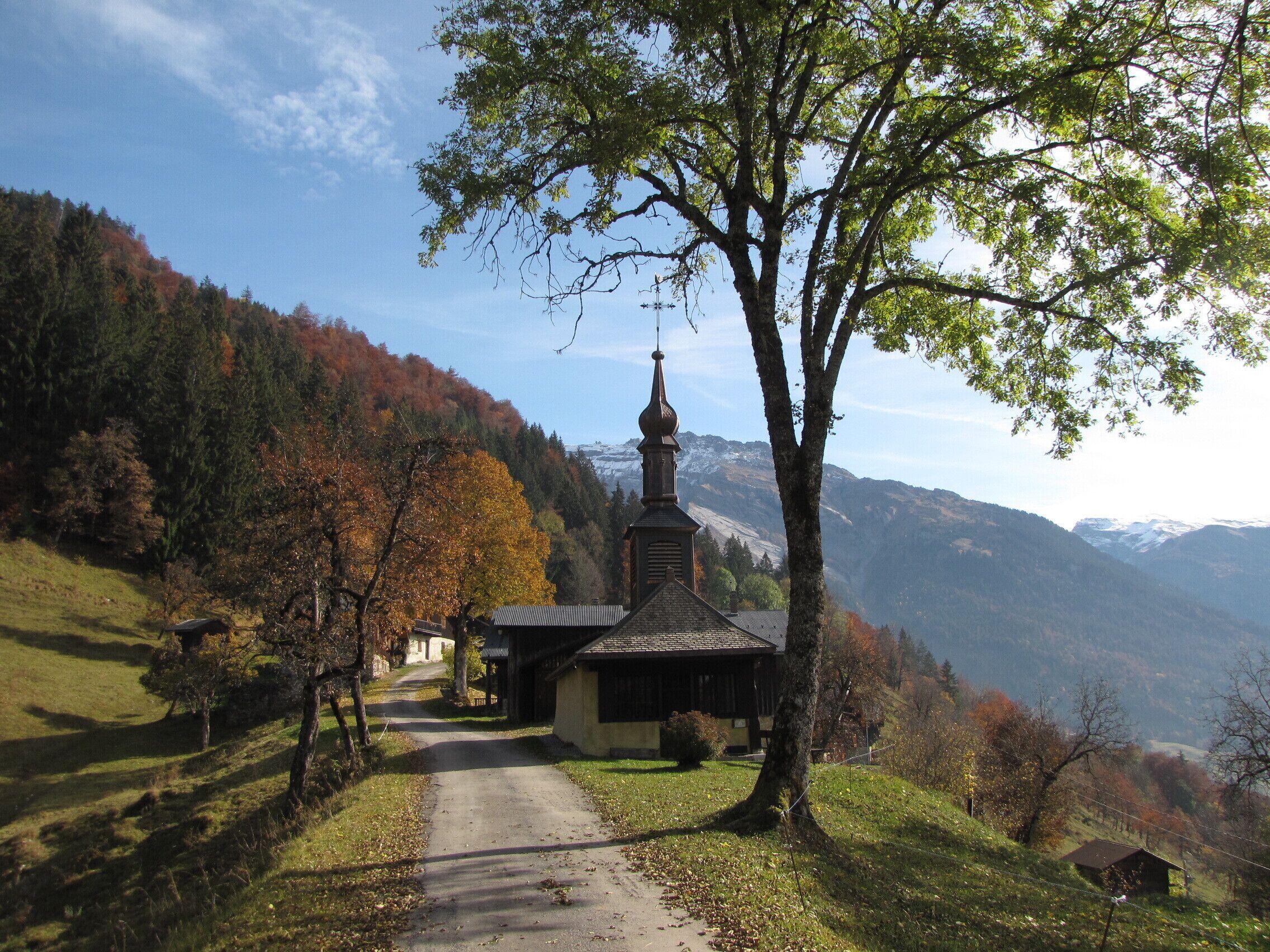 Església rural amb un campanar, envoltada de fullatge d'hivern i muntanyes al fons.