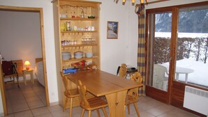 Ein Essbereich mit Holztisch und Stühlen, einem Buffetkorpus mit Geschirr und Blick auf eine verschneite Landschaft durch eine Glastür.