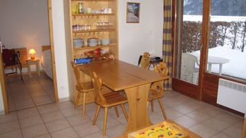 Een eetruimte met een houten tafel en stoelen, een buffetkast met servies, en zicht op een besneeerd landschap door een glazen deur.