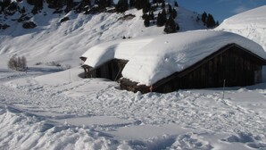 Ein verschneites Holzhäuschen mit geneigtem Dach, umgeben von verschneiter Landschaft und einem bewaldeten Berg im Hintergrund.
