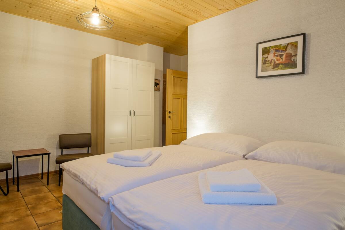 A hotel room with two beds, a wooden ceiling, a white wardrobe, a small table, and a chair.