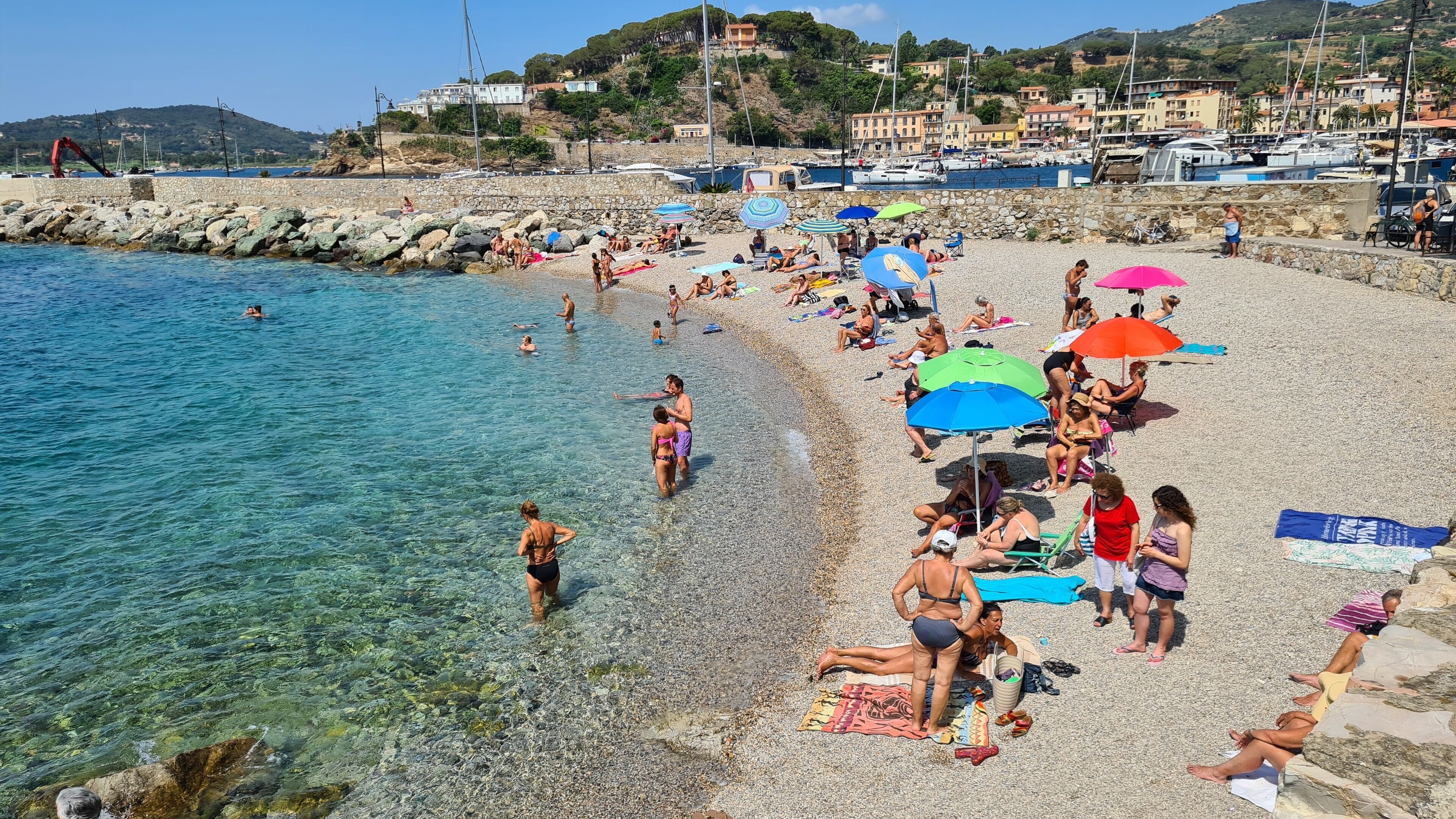 Ein überfüllter Strand mit klarem Wasser, Menschen, die sich sonnen, und Booten im Hintergrund.