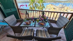 A balcony with wicker furniture, a table set with breakfast items, and a view of a beach and boats. - Blue harbor lovely apartment with terrace above the beach. (Porto Azzurro)