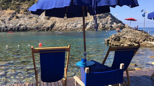 Two blue beach chairs with a table and a blue umbrella on a wooden deck overlooking a rocky shoreline with clear water and boats.