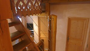 Eine Holztreppe mit dekorativer Geländer führt zu einem Zimmer im zweiten Stock. Im Zimmer ist eine Holztür und ein kleines Fenster zu sehen.