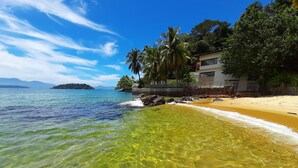 A beach with clear water, a sandy shore, and a building with a stone wall and a fence. - Wonderful house by the water in Angra dos Reis. (Angra dos Reis)