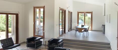 Un vaste espace de vie comprenant un canapé moderne, une table basse et une table à manger avec des chaises. De grandes fenêtres laissent entrer la lumière naturelle et offrent une vue sur l’extérieur.