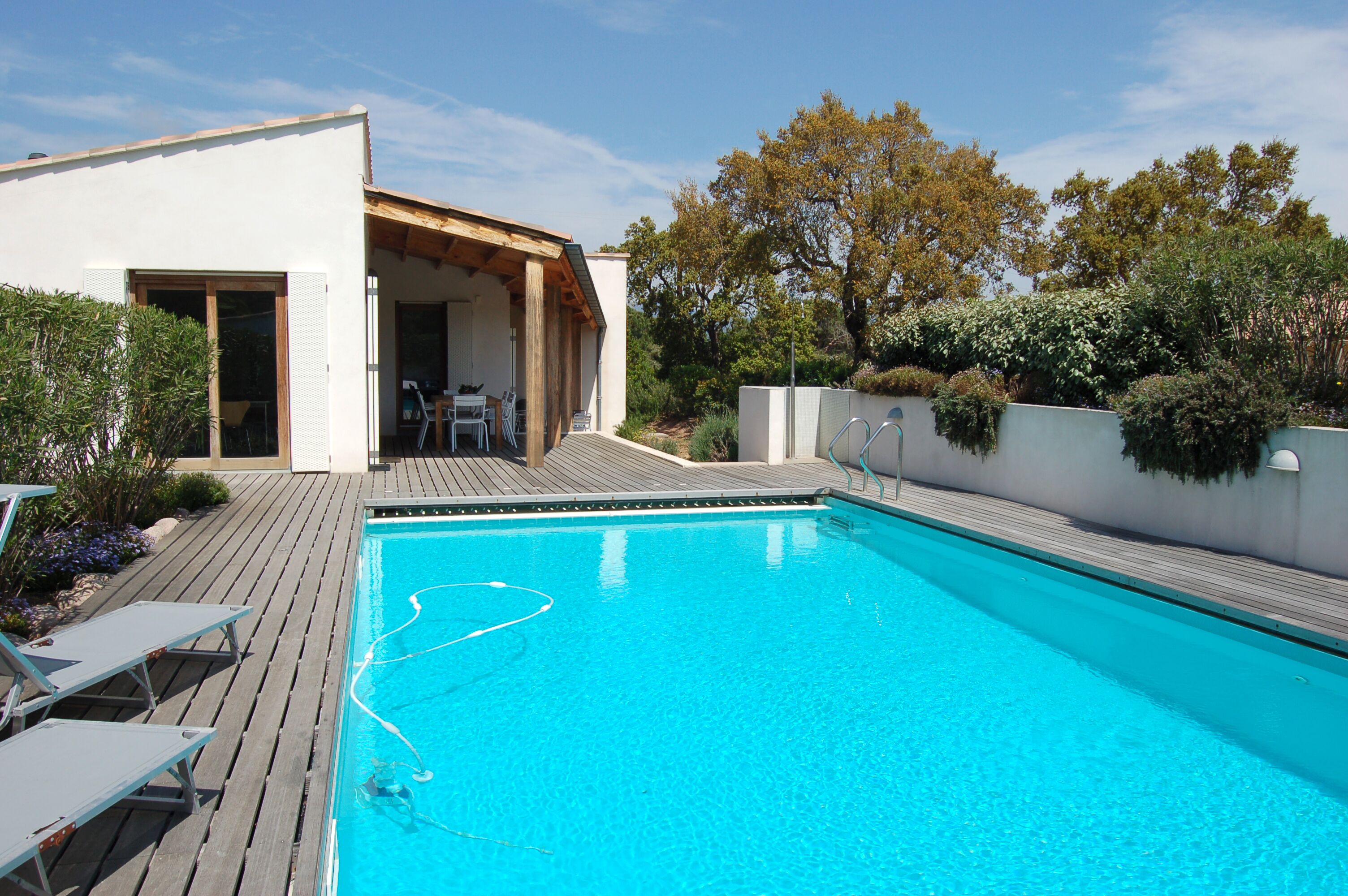 A modern outdoor swimming pool with a wooden deck, a covered seating area, and lush greenery.