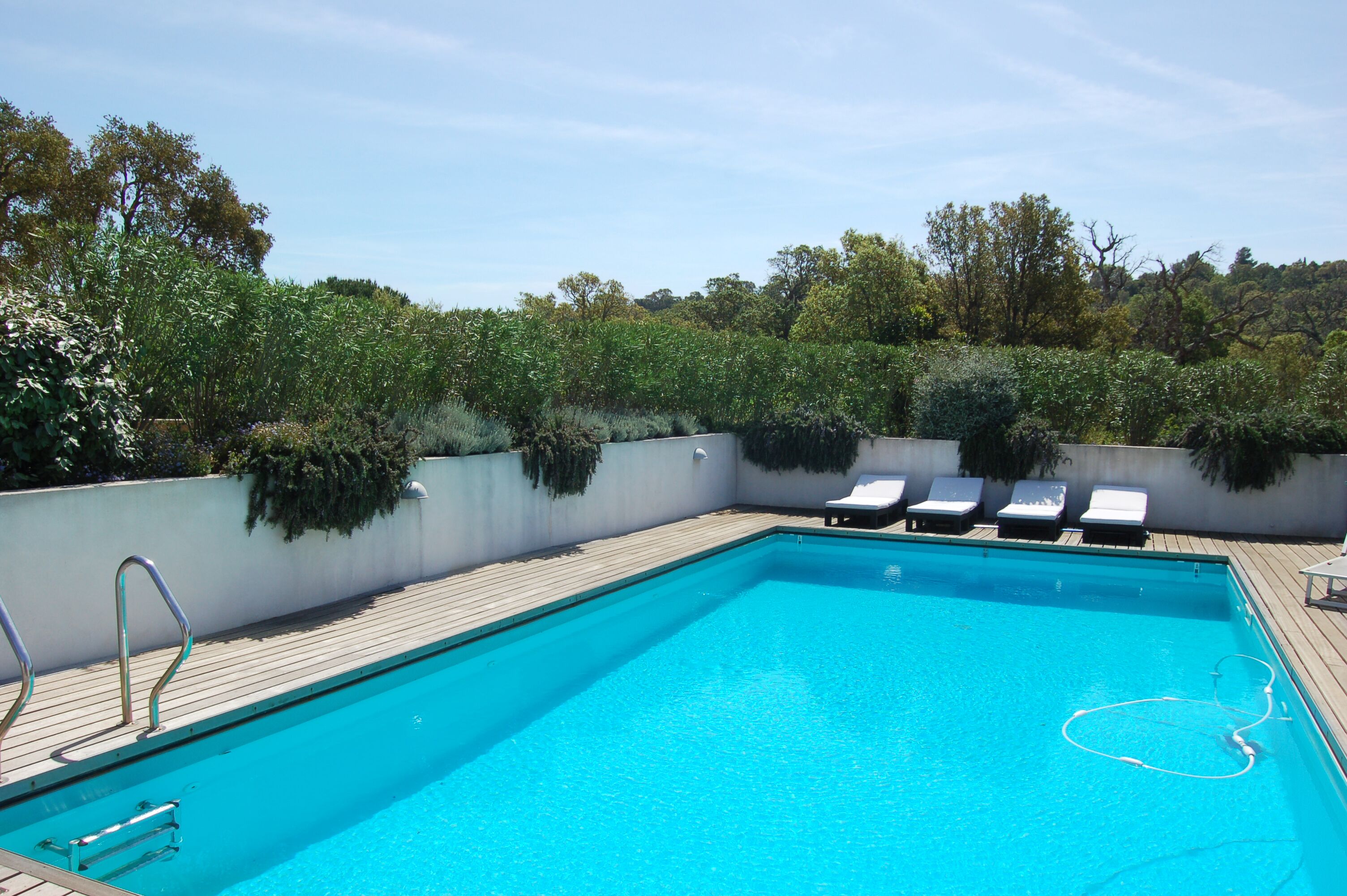 A rectangular swimming pool with a wooden deck and metal railings. There are four lounge chairs aligned along the pool's edge.