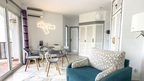 Een moderne woonkamer met een blauwgroene bank, een glazen eettafel met stoelen en een sierkussen met een patroon.