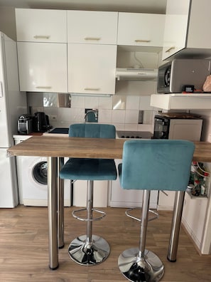 Une cuisine moderne avec des meubles blancs, une table en bois, des tabourets de bar turquoise et un lave-linge.