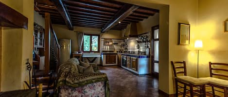 Amplia sala de estar con sofá, sillas de madera, una cocina al fondo y una escalera que sube.
