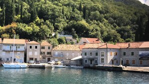 Un village côtier aux bâtiments en pierre, un port de plaisance avec des bateaux, et un paysage montagneux en arrière-plan.