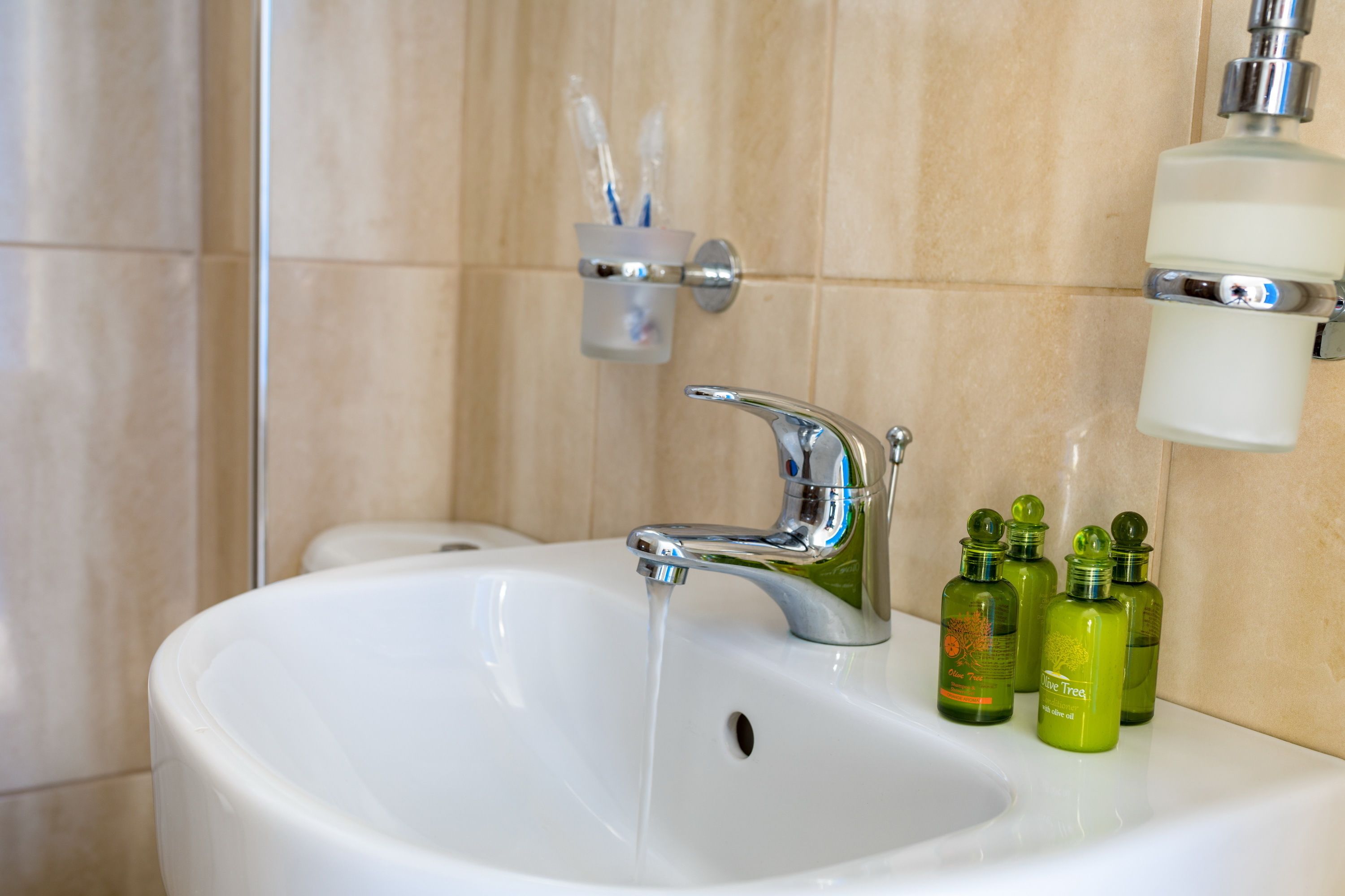 Un lavabo blanc dans la salle de bain, avec un robinet chromé, une tasse remplie de brosses à dents et trois bouteilles d’huile d’olive posées sur le comptoir.