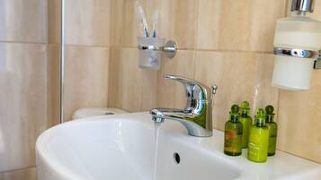 Un lavabo blanc dans la salle de bain, avec un robinet chromé, une tasse remplie de brosses à dents et trois bouteilles d’huile d’olive posées sur le comptoir.