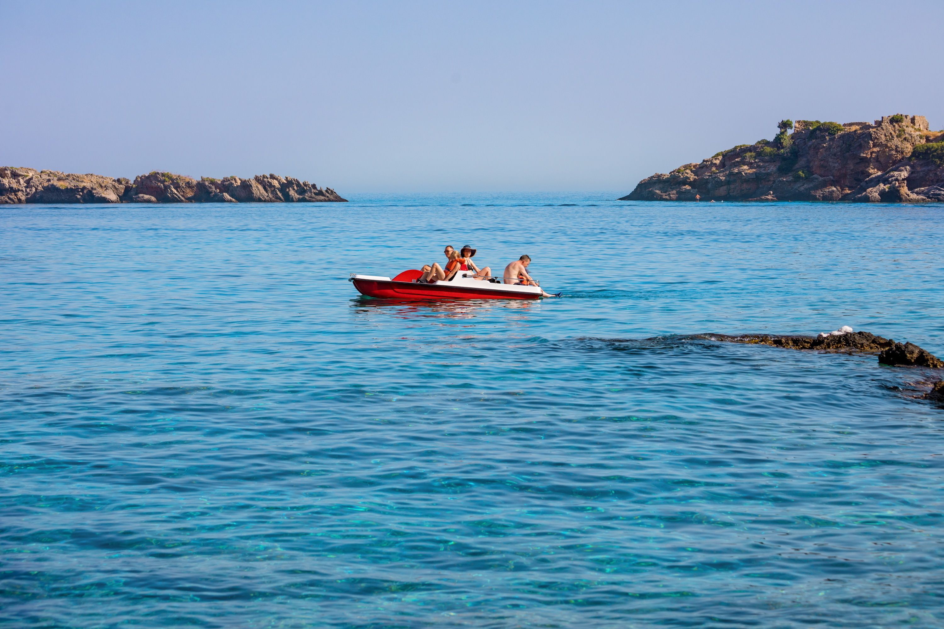 A small red boat with people on a calm sea, rocky islands in the background.