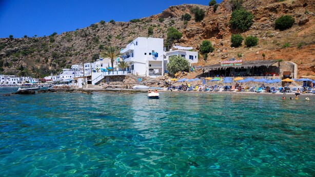 A coastal town with clear blue waters, a white building with balconies, and a beach area with umbrellas and people. - John Akroyiali rooms (Sfakia)