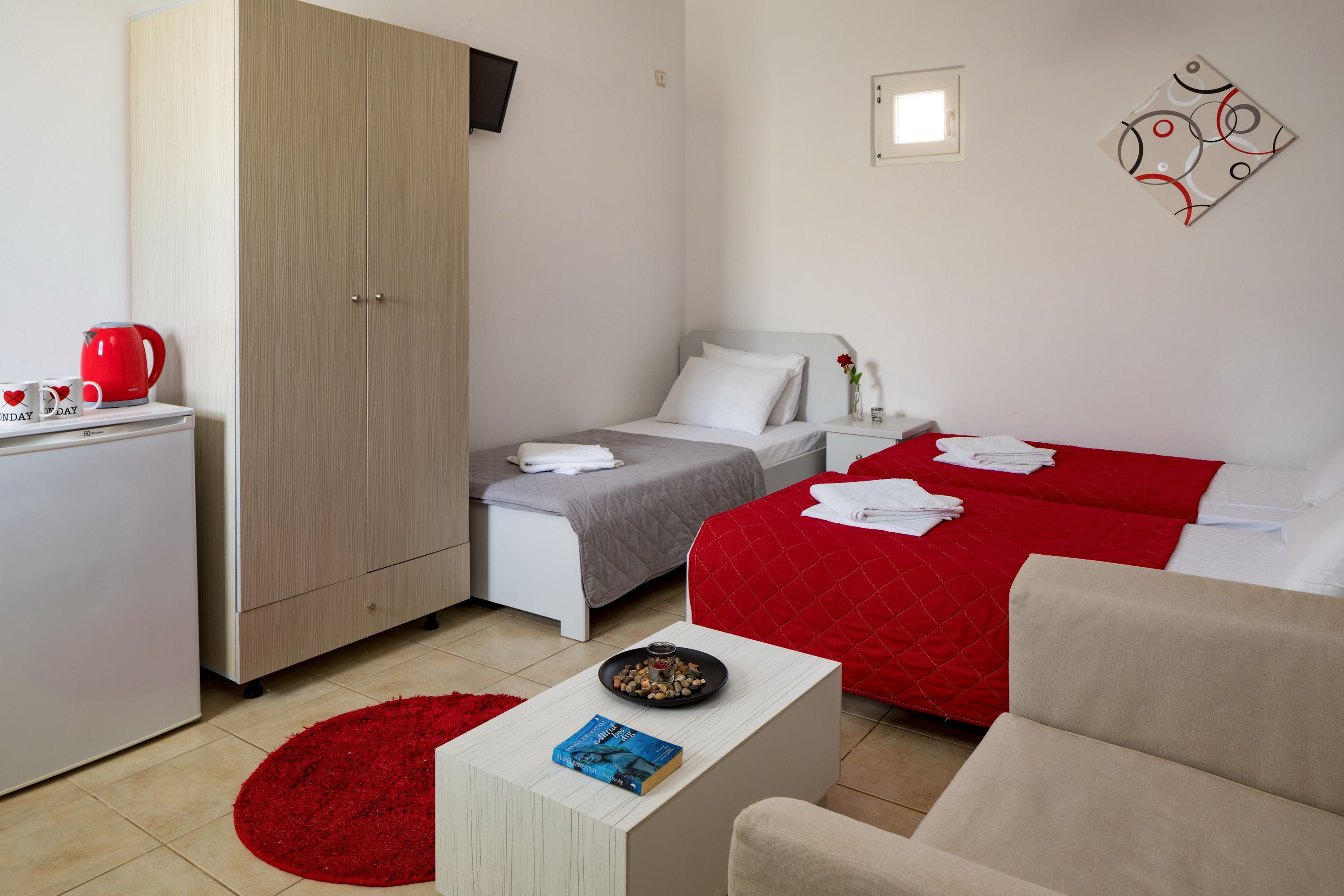 A hotel room with a bed, a desk with a red kettle, a bookshelf, a red rug, and a wall art piece.