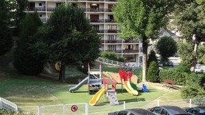 A playground with slides and a fenced area, surrounded by trees and a multi-story building in the background. - Perfect  location quiet surroundings  spacious comfortable & functional  (Brides-Les-Bains)