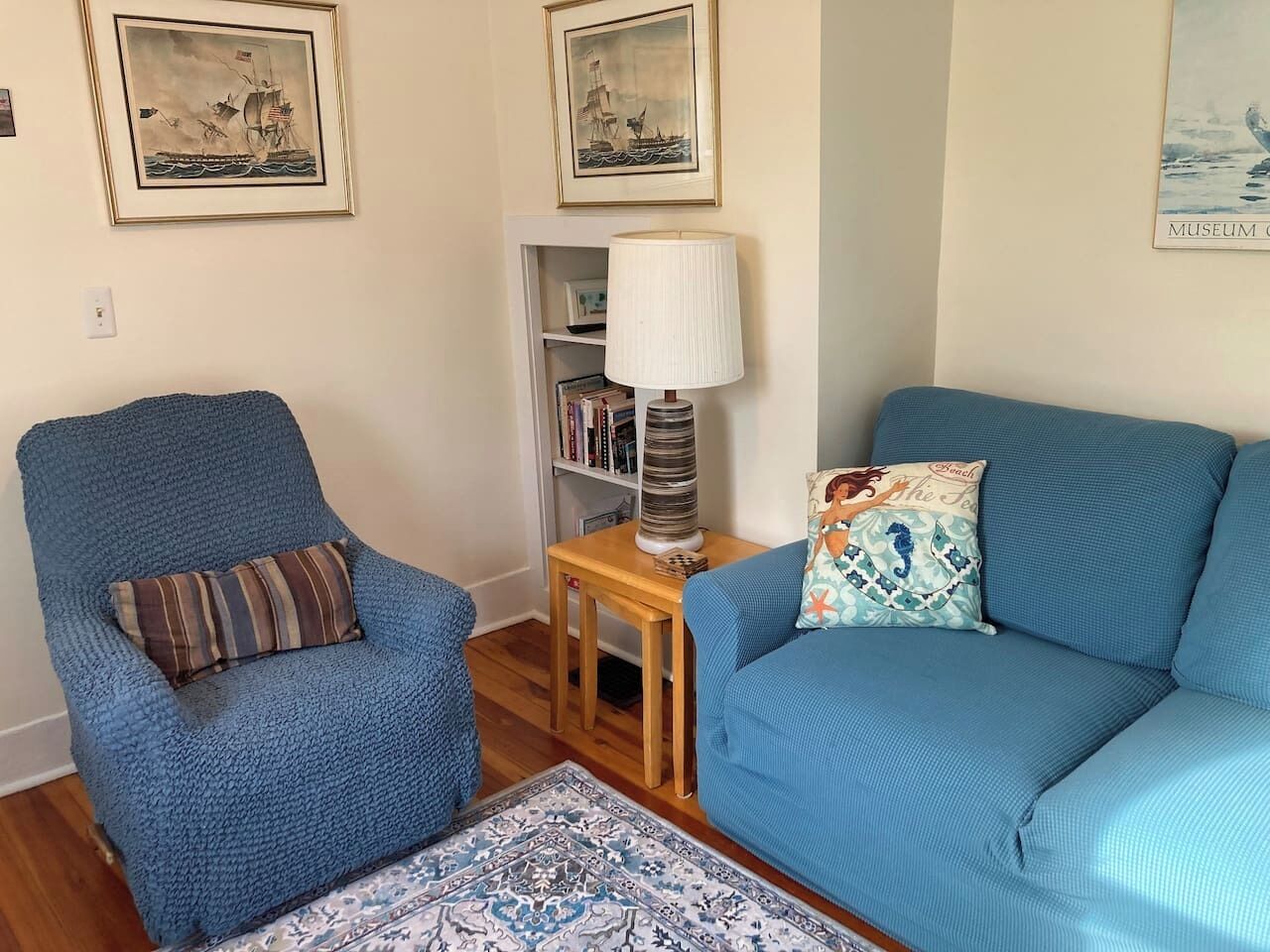 A living room with a blue armchair, a blue sofa, a small wooden side table with a lamp, and a bookshelf.