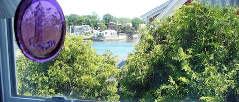 A view through a window with a curtain, overlooking a body of water, greenery, and buildings in the distance.
